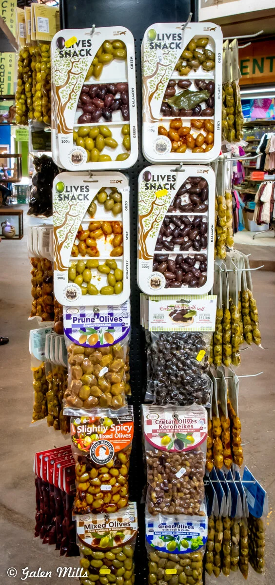 Variety of packaged olives on display, including green, spicy, and Kalamon olives, in a market setting.