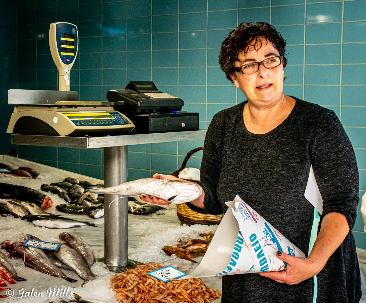 Woman holding a fish at a seafood market counter with weighing scales and various fish on ice.