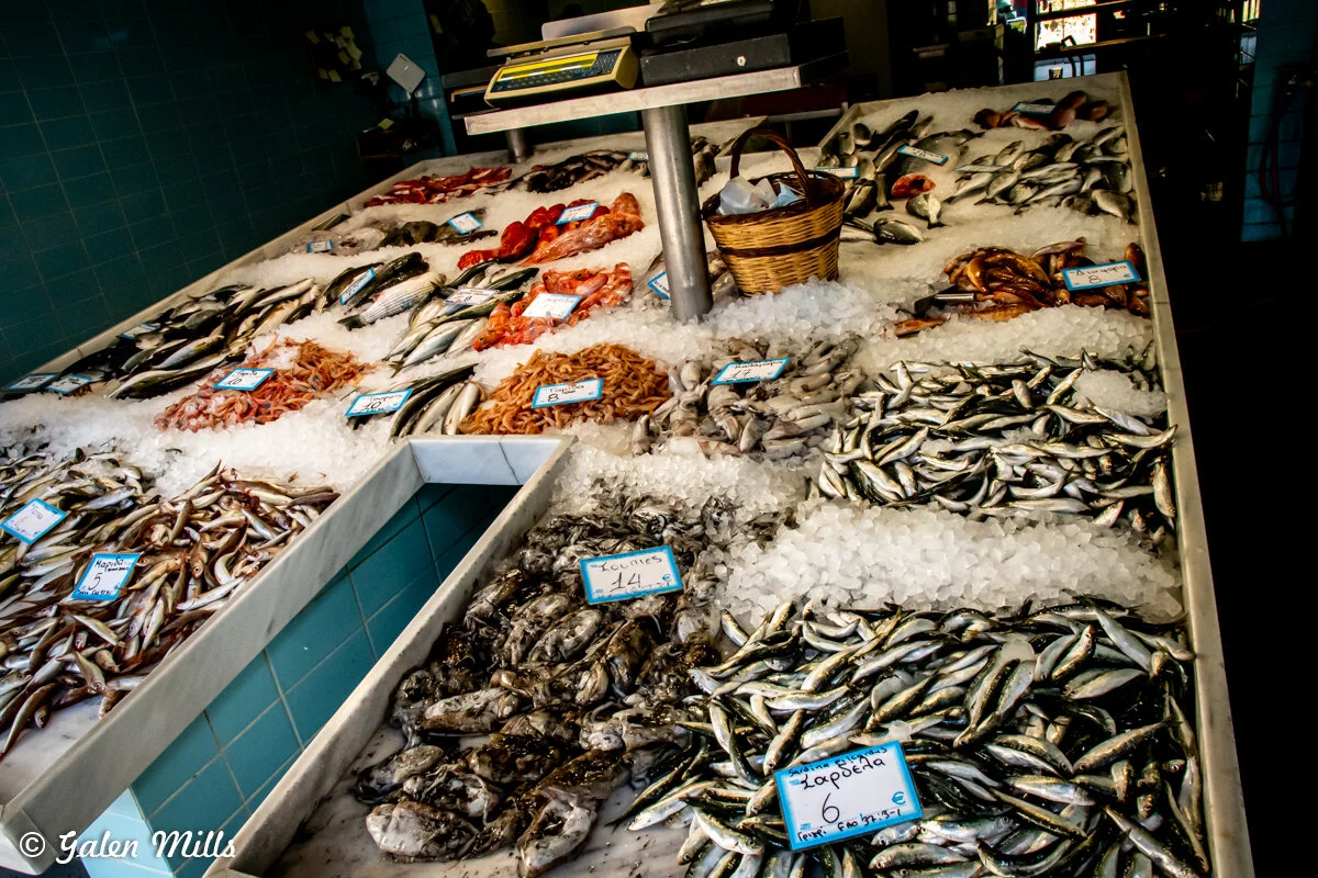Fish market display with various types of fish and seafood on ice, including small fish, shrimp, and other seafood items, with price tags and a basket on the counter.