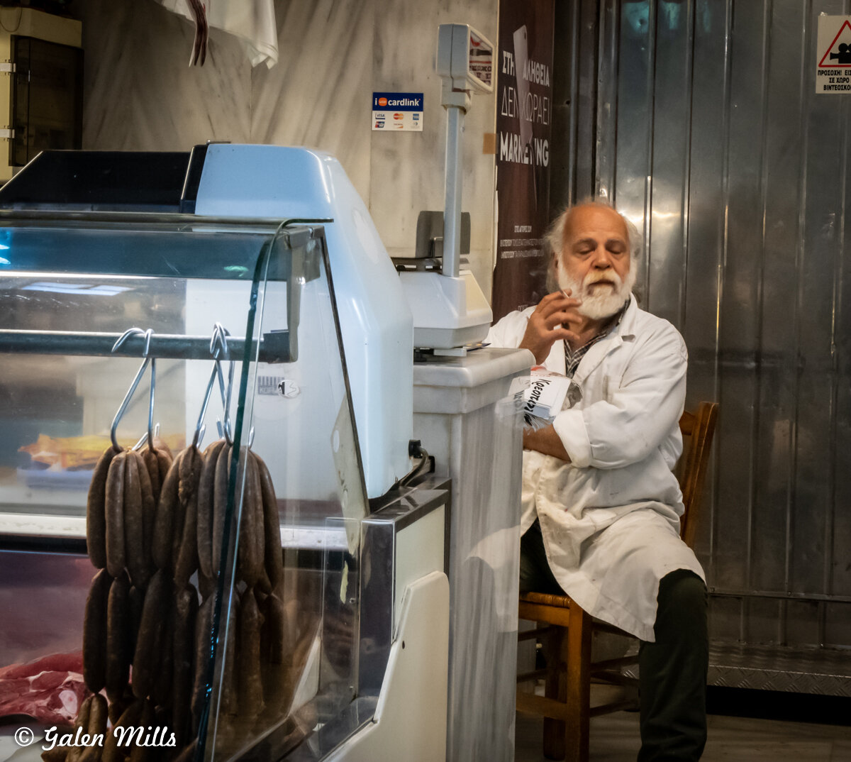 Butcher sitting in shop next to meat display case with hanging sausages.