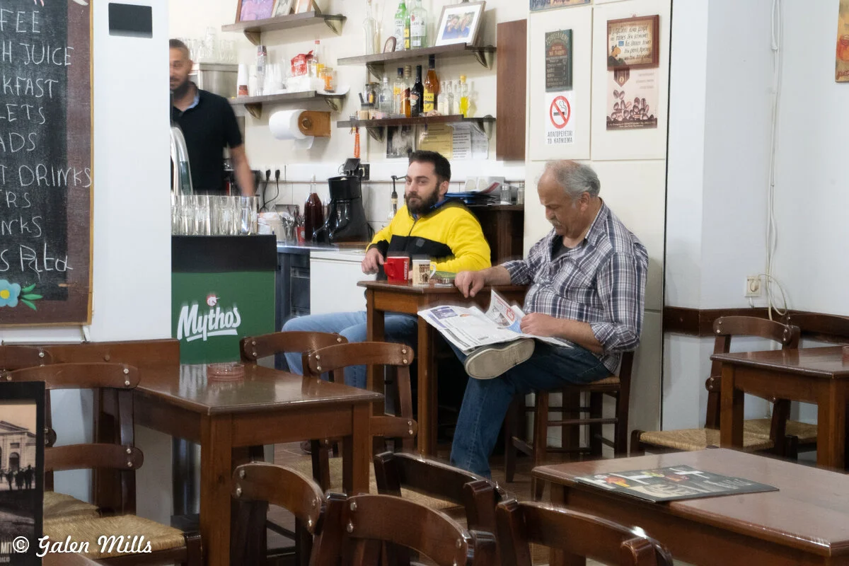 Two men sitting in a cafe, with one reading a newspaper and the other drinking from a mug. The cafe has wooden tables and chairs, a menu board, and shelves with bottles and glassware. Another person is visible behind the counter.