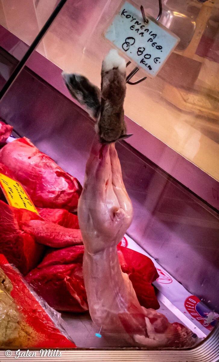 A skinned rabbit hanging in a butcher's display case with a label showing the price per kilogram. Fresh cuts of red meat are visible in the background.