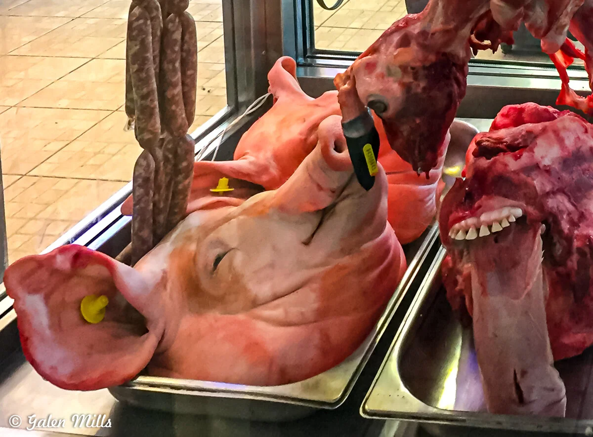 A butcher shop display featuring a pig's head, sausages, and meat cuts on trays.