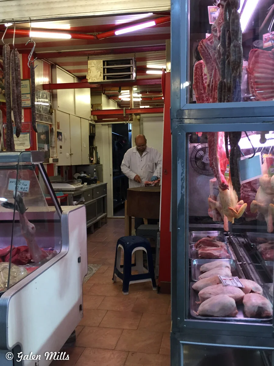 Interior of a butcher shop with meat displayed in glass cases, sausages hanging, and a butcher working in a white coat.
