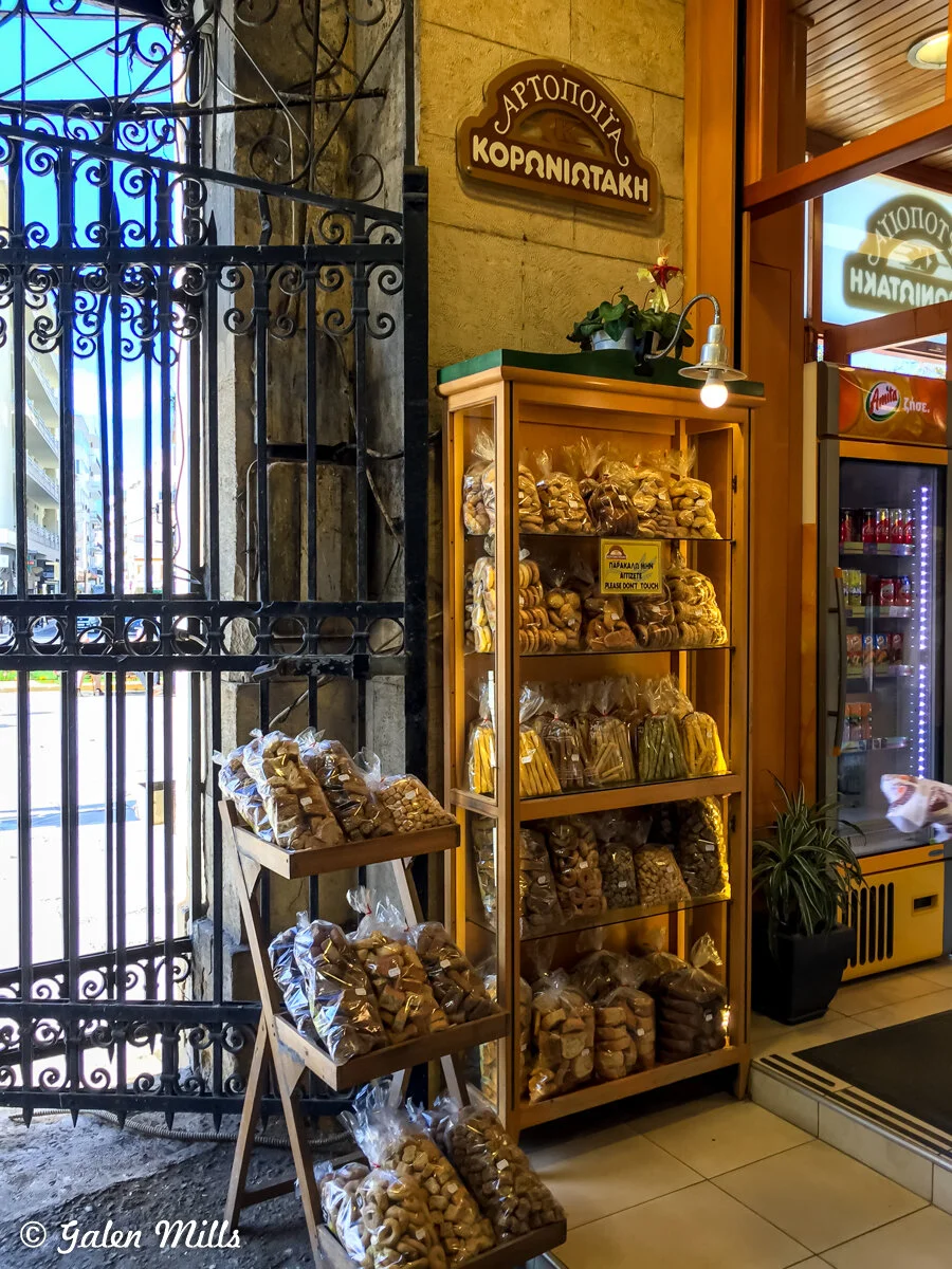 Bakery entrance displaying shelves of packaged baked goods, with a decorative iron gate on the left and a sign above indicating it's a bakery.