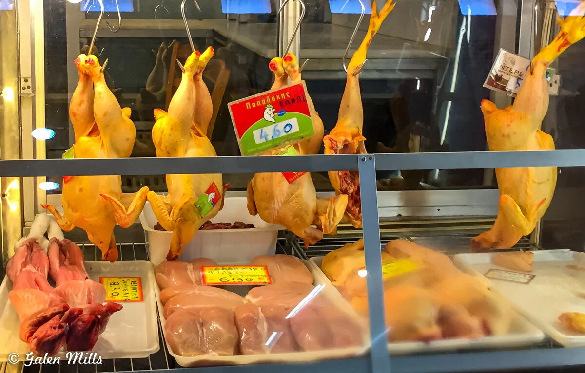 A display of raw poultry and meat in a market, featuring whole chickens hanging upside down and various cuts of chicken meat on trays, with price tags attached.