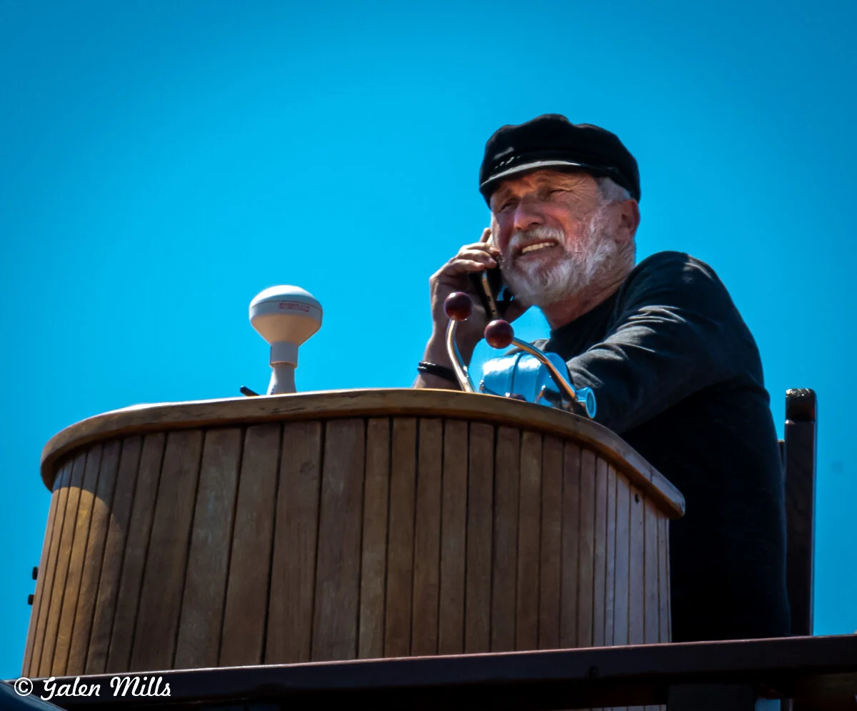 Man with a beard and cap speaking on a phone while standing at a wooden ship's helm with blue sky background.