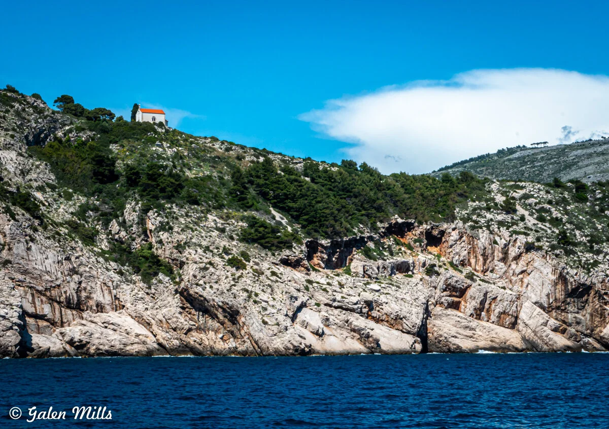 Coastal rocky hillside with a small white chapel and a red roof on top, surrounded by greenery under a blue sky.