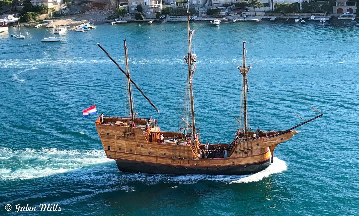 Wooden sailing ship on blue water, nearby shore with boats and houses.