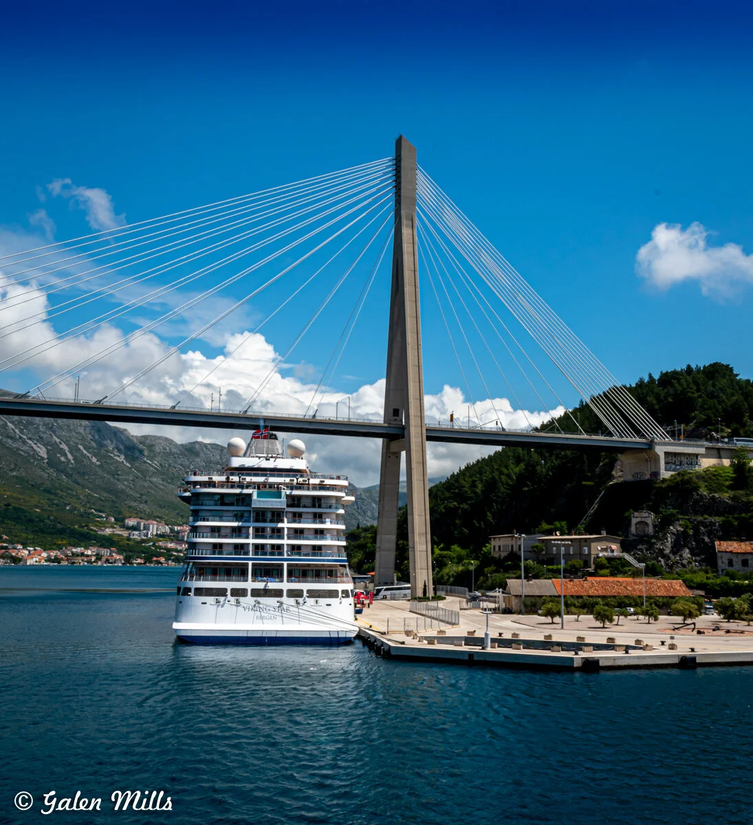 Cruise ship docked near a cable-stayed bridge with mountainous landscape and clear blue sky in the background.