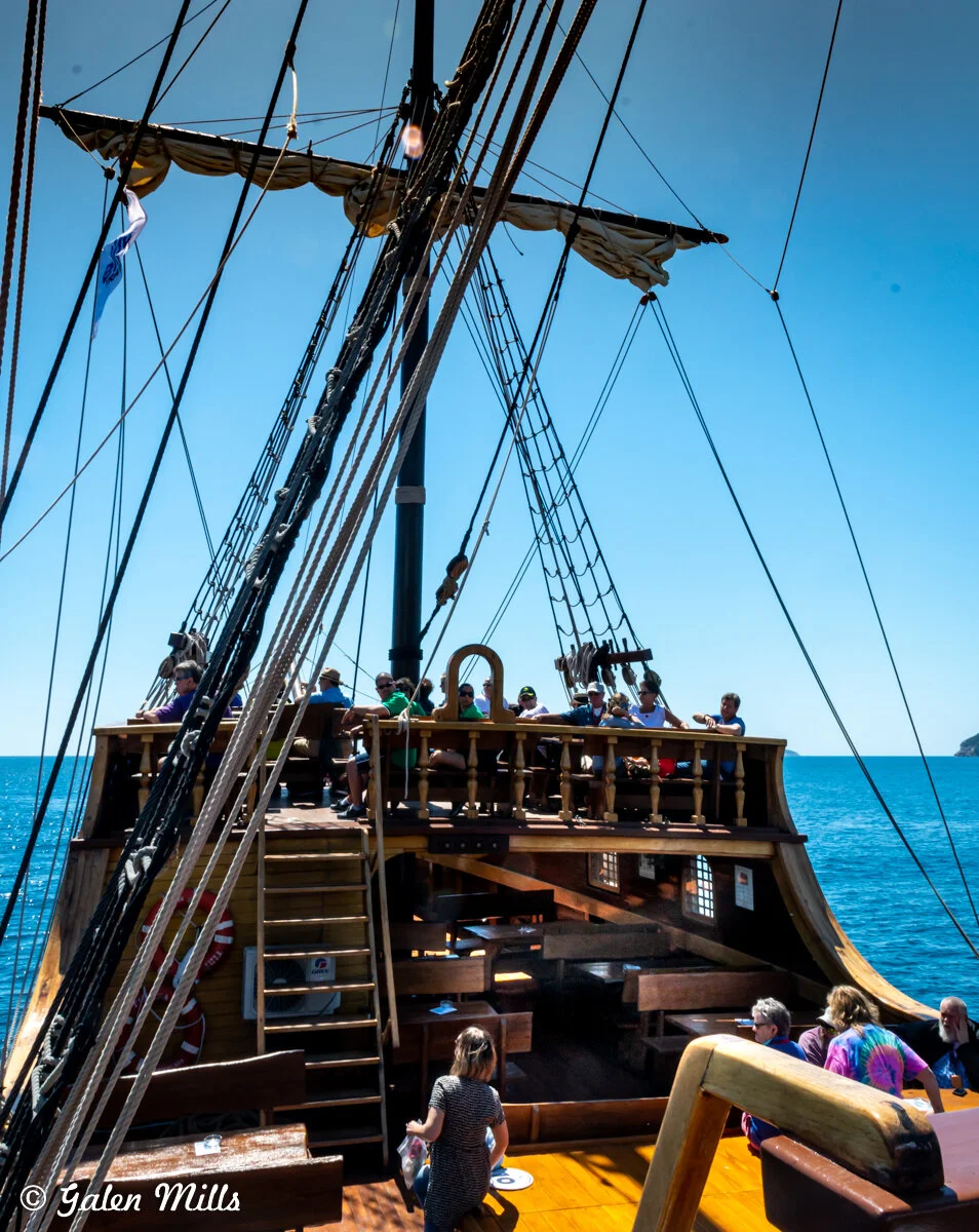 People on the deck of a wooden sailing ship with ropes and masts visible, under a clear blue sky.