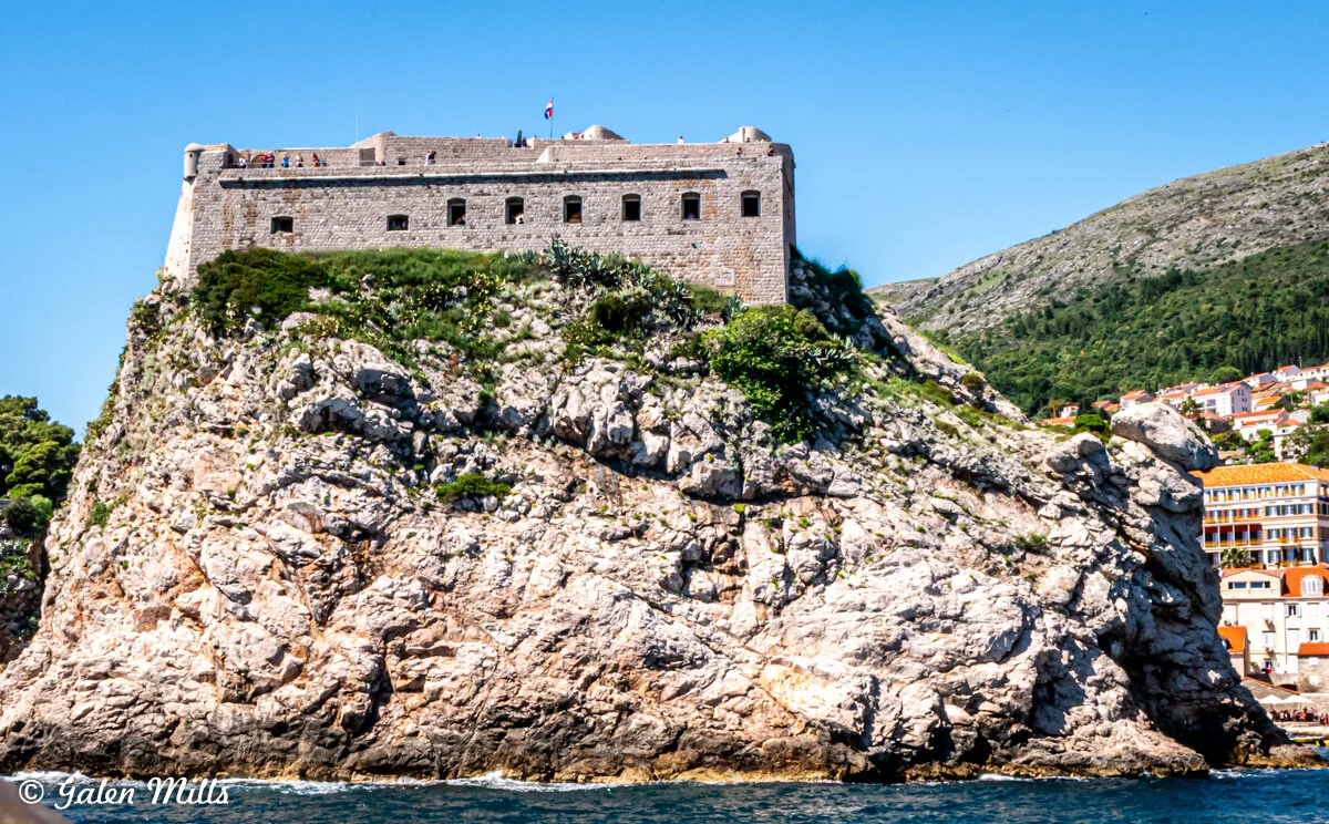 Fortress atop a rocky cliff by the sea with a view of nearby buildings and hills.