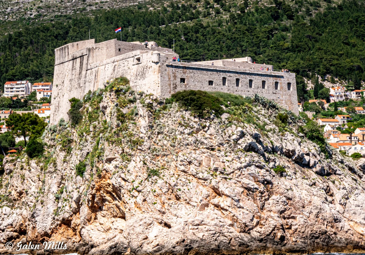 Fortification on rocky cliff with vegetation, surrounded by green hills and buildings with red roofs.
