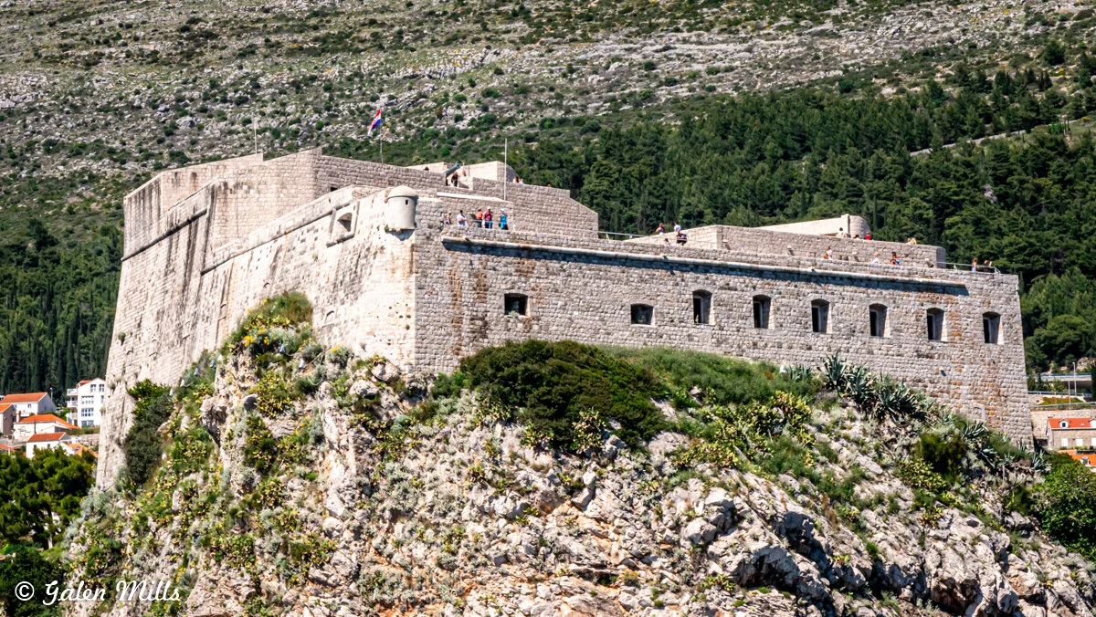A large stone fortress on a rocky hill with people on top, surrounded by greenery and shrubs, with a flag visible in the background.
