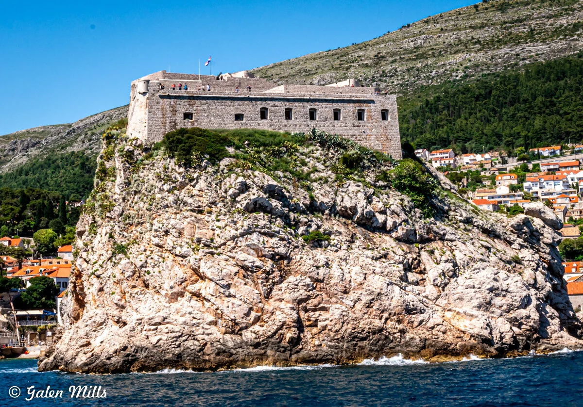 Fortress atop a rocky cliff by the sea, surrounded by greenery and adjacent to a town with orange-roofed buildings.