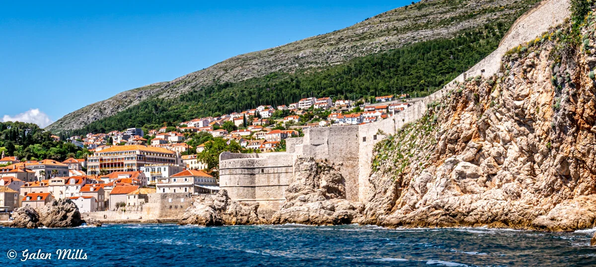 View of Dubrovnik Old Town city walls and orange-roofed buildings with rocky coastline and hillside in background.