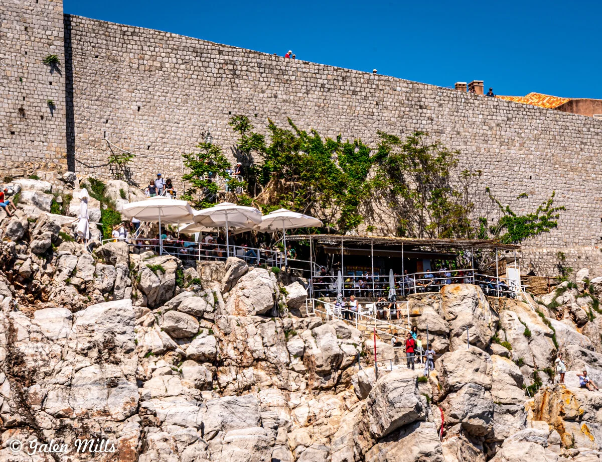 Outdoor cafe on rocky cliff with umbrellas, people sitting, and large stone wall in background under clear blue sky.