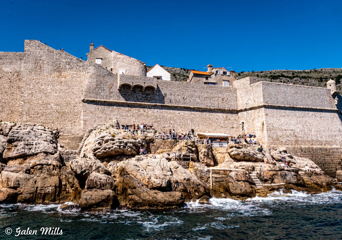 View of historic stone fortifications on a rocky coastline with people gathered on the rocks and a clear blue sky.