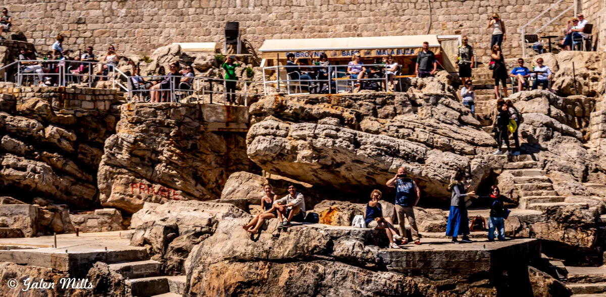 People sitting and standing on rocky cliffs near a cafe with a stone wall backdrop, enjoying the outdoors.