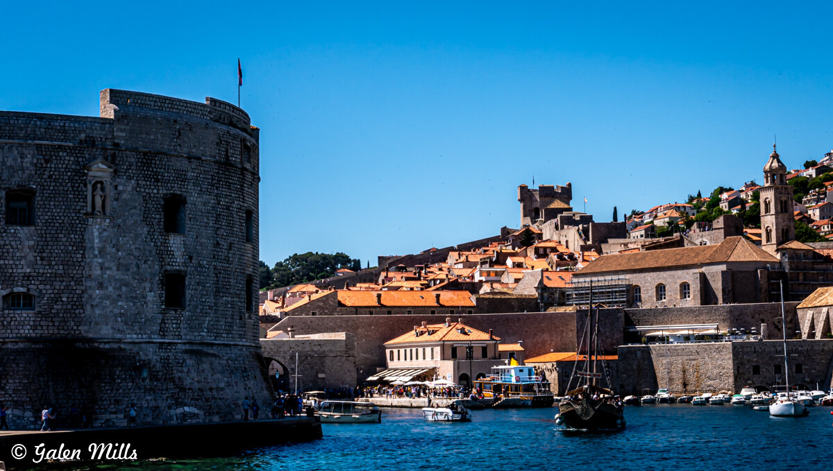View of Dubrovnik's historic city walls and harbor with boats, old stone buildings, and clear blue sky.