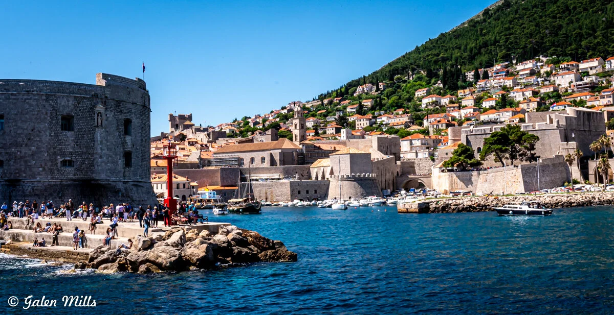 Scenic view of Dubrovnik Old Town with historic stone walls, boats on the Adriatic Sea, and hillside houses under a clear blue sky.