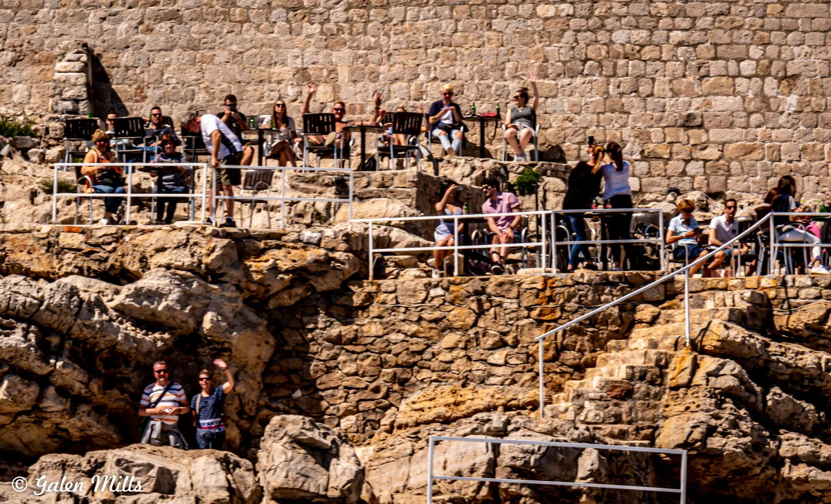 People sitting at outdoor cafe on rocky cliff, with stone wall backdrop.