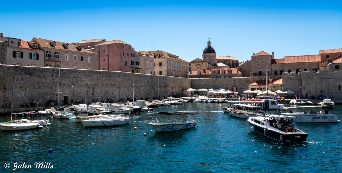 Boats docked in a harbor with historic stone buildings and a domed cathedral in the background, under a clear blue sky.