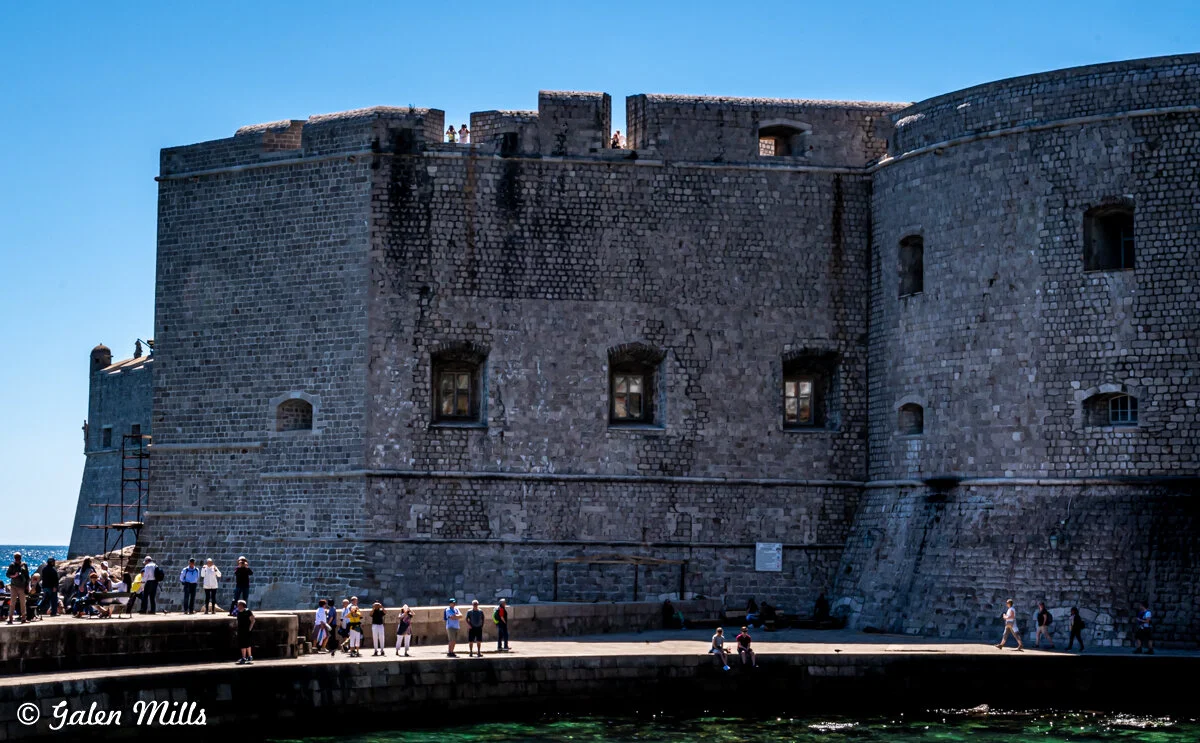 Large stone fortress wall with windows, surrounded by people walking and standing on a waterfront path, with a clear blue sky in the background.
