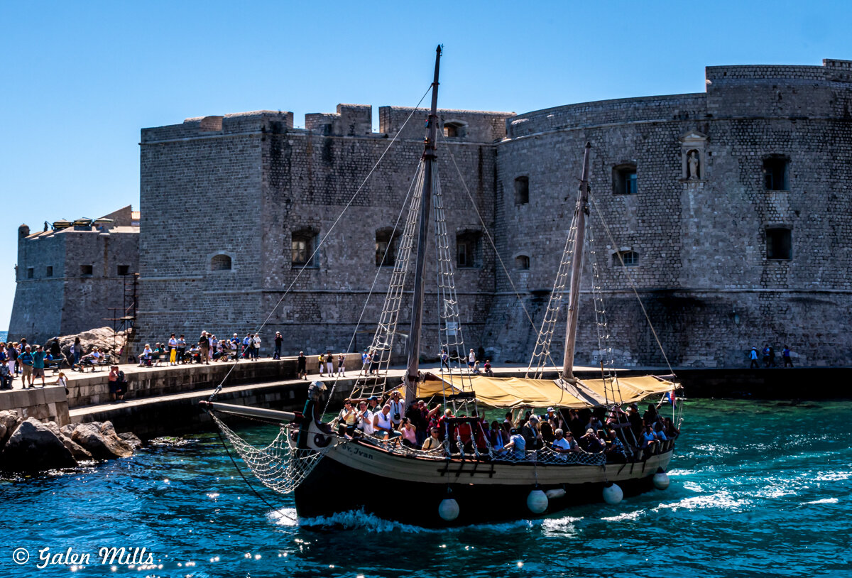 Tourist boat with masts sailing near historical stone fortress in Dubrovnik, Croatia, on a sunny day.