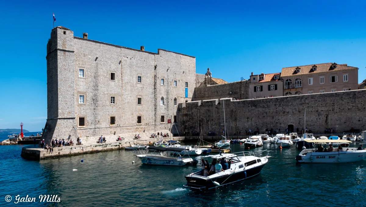 View of Dubrovnik's harbor with old city walls, boats docked in the water, and a clear blue sky.