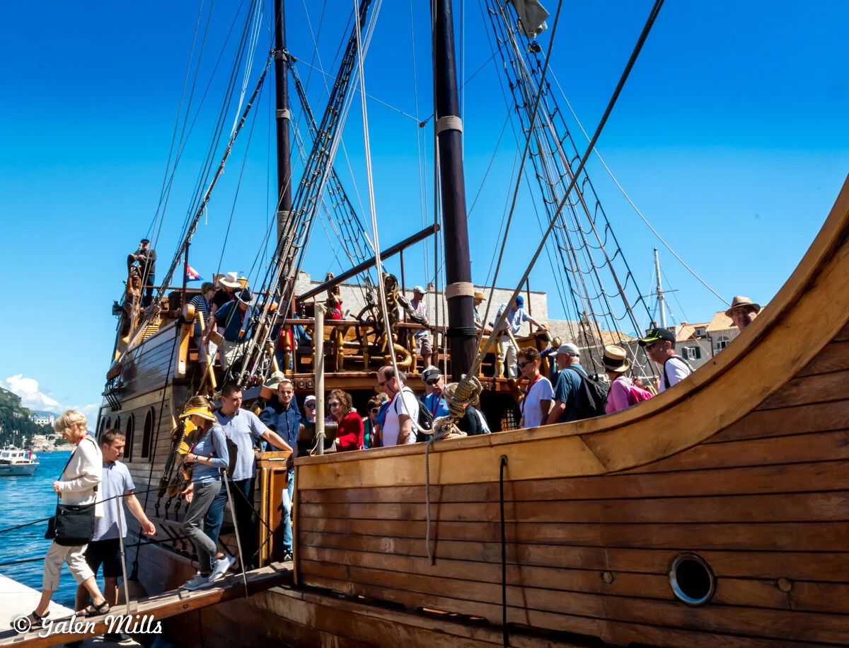 Large wooden sailing ship with visitors on deck in a scenic harbor.