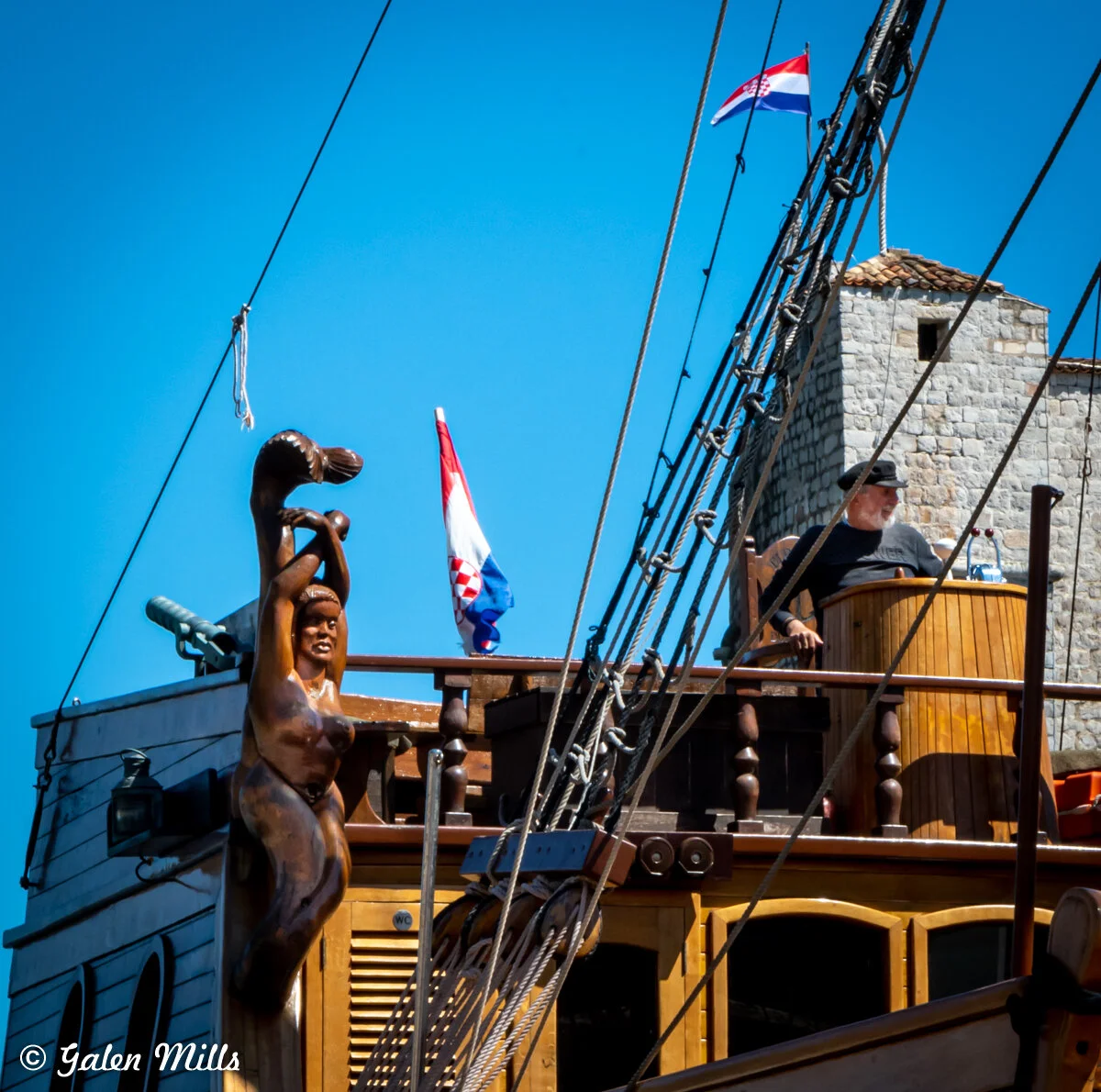 Wooden figurehead of a mermaid on a historic ship's prow, with a man on deck and a Croatian flag, against a blue sky and stone building.