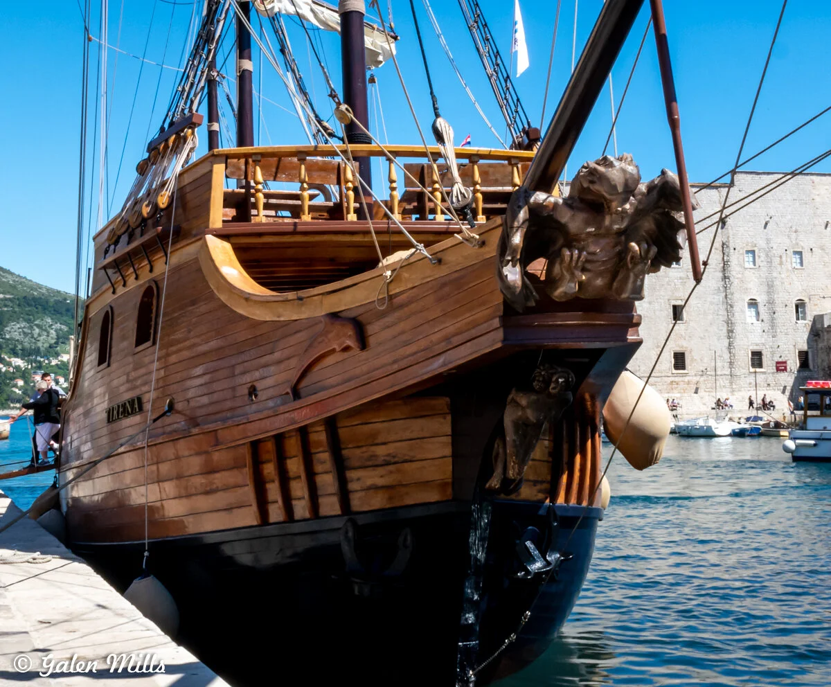 Close-up of a wooden sailing ship with a decorative figurehead, docked at a harbor with buildings and hills in the background.