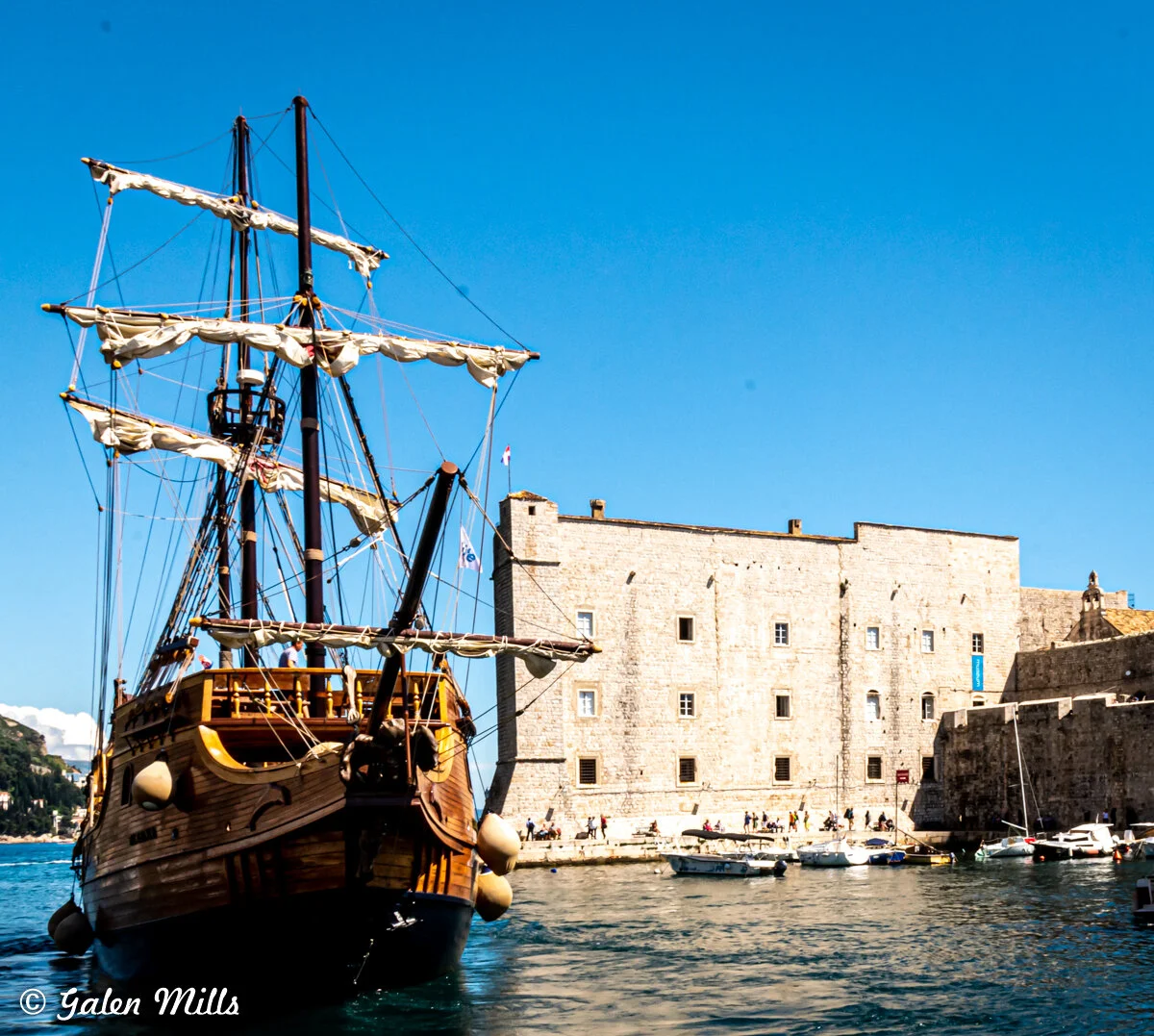 Historic sailing ship in harbor near stone building on clear day