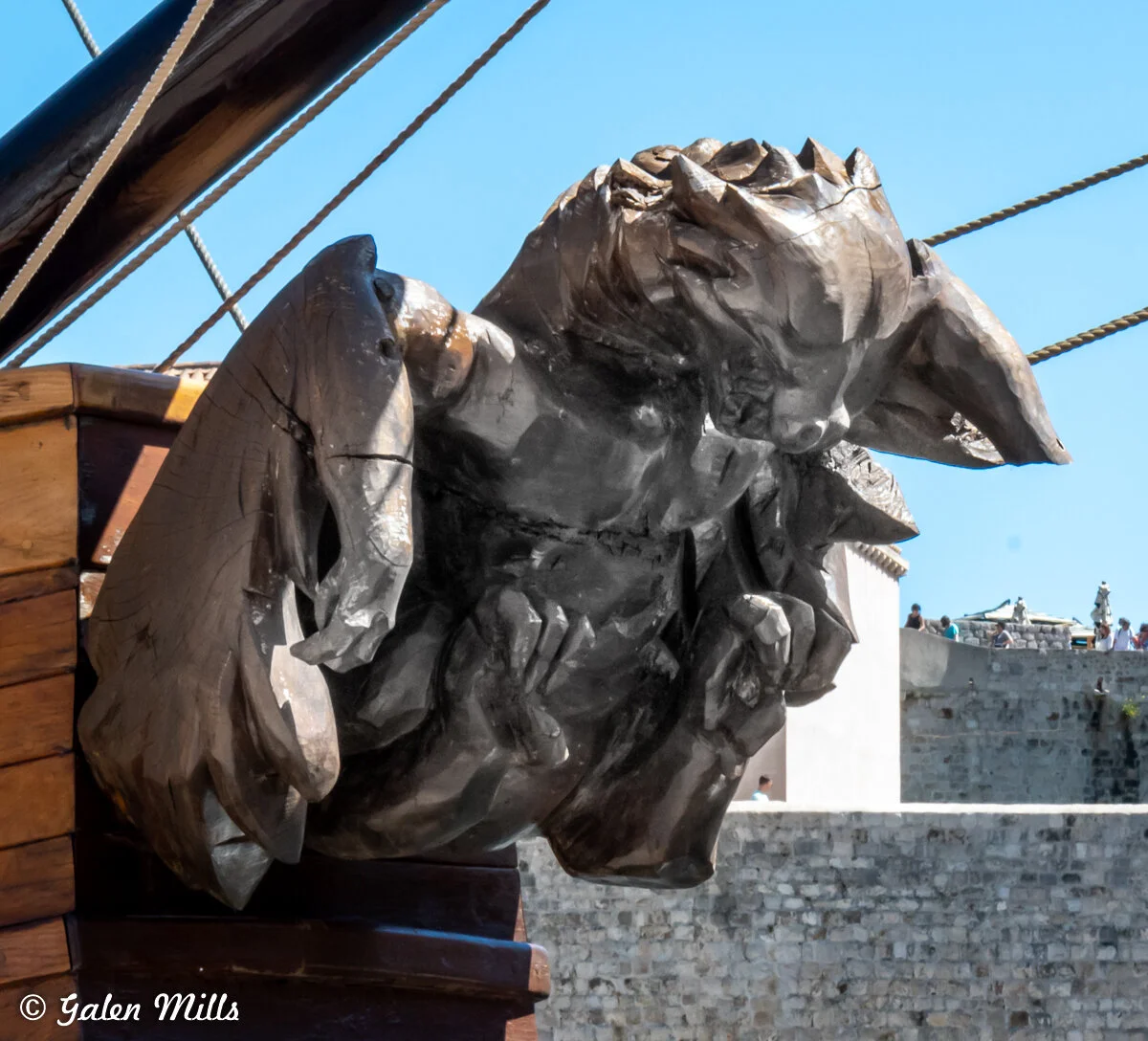 Wooden gargoyle figurehead on a ship with ornate carvings, set against a blue sky and building in the background.