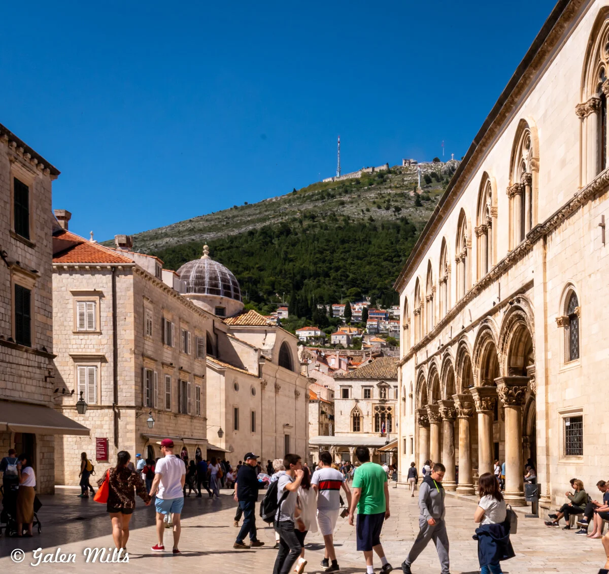 Tourists walking in a historic European city square, surrounded by stone buildings with arches and a dome, with a hill and tower in the background, under a clear blue sky.