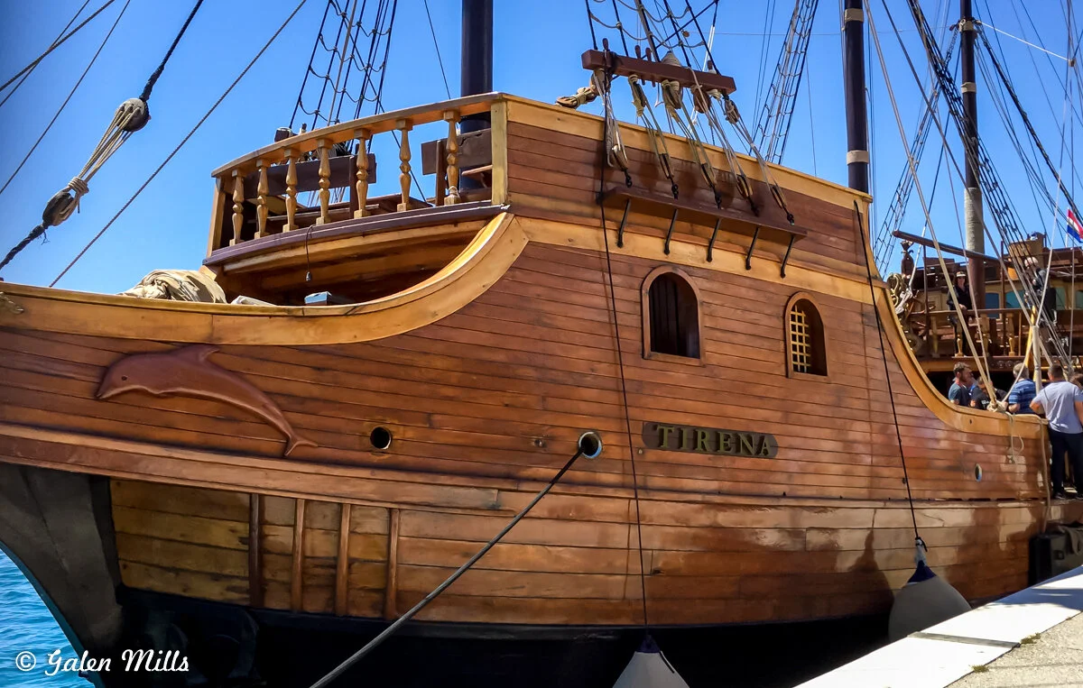 Wooden sailboat named "Tirena" docked at a pier with blue sky background.