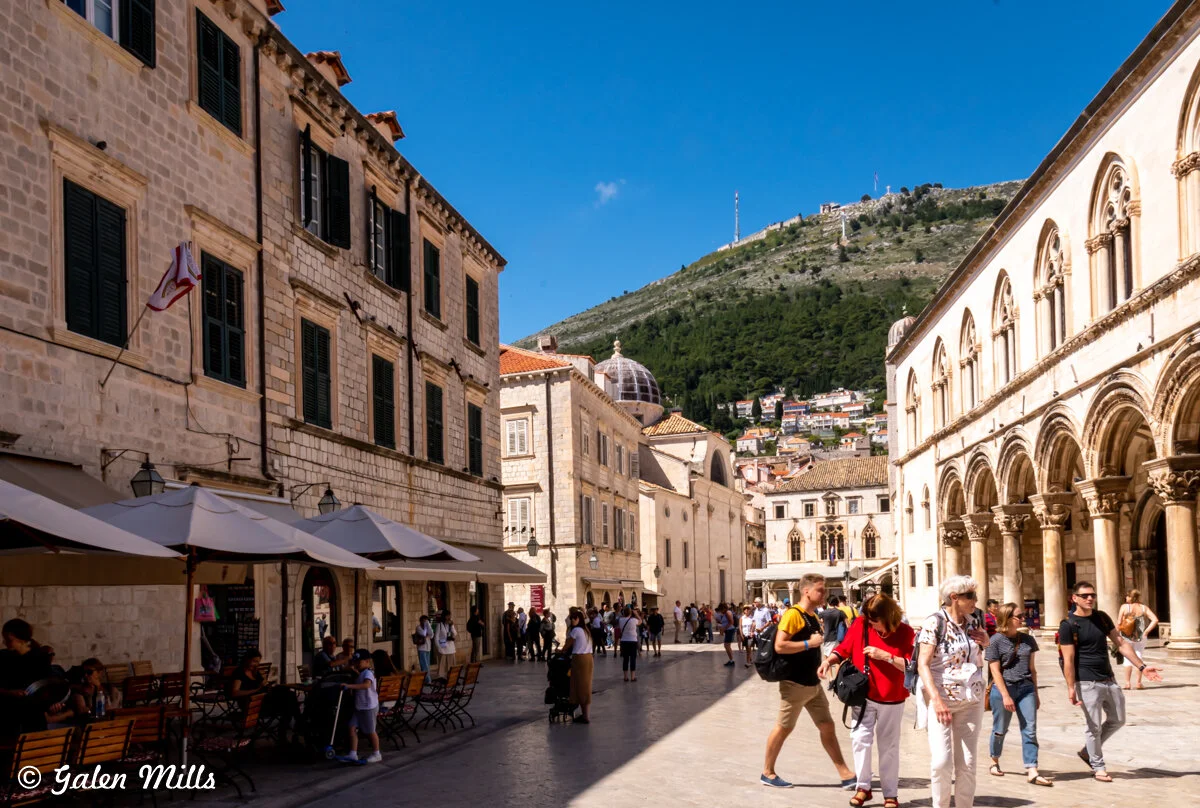 People walking in a sunlit plaza in Dubrovnik, Croatia, surrounded by historic stone buildings and outdoor cafes, with a hilly backdrop.