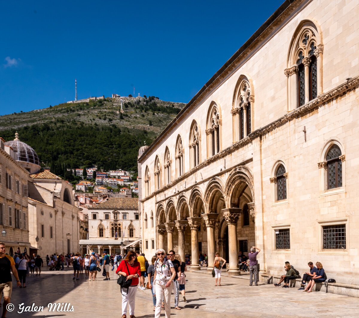 Tourists walking and sitting at Rector's Palace in Dubrovnik, Croatia, featuring Venetian Gothic architecture, with hills and cityscape in the background.