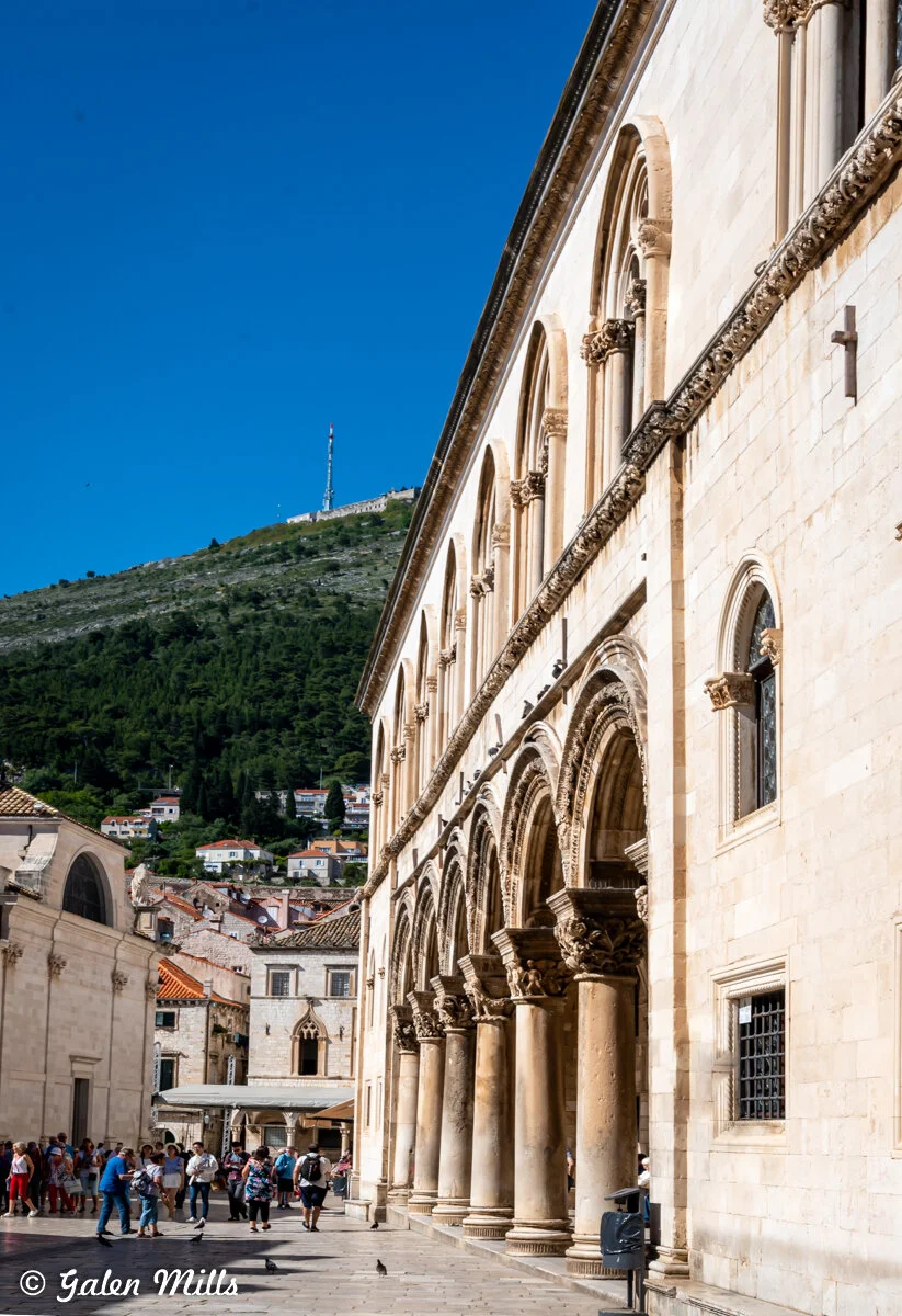 A historic building with arches and columns in Dubrovnik, Croatia, under a clear blue sky. A hill with greenery and a telecommunications tower is visible in the background. People are walking and gathering in the foreground.