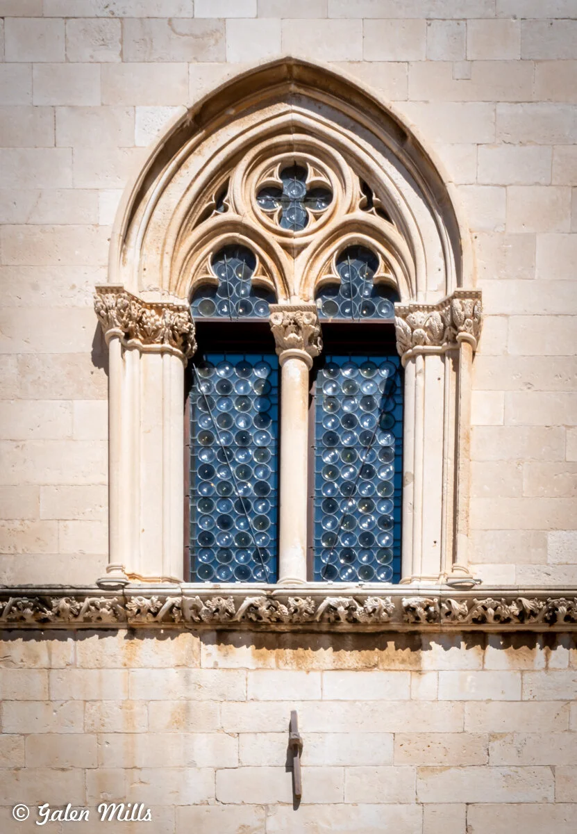 Gothic-style arched stone window with ornate columns and circular patterned glass panes on a beige brick building façade.