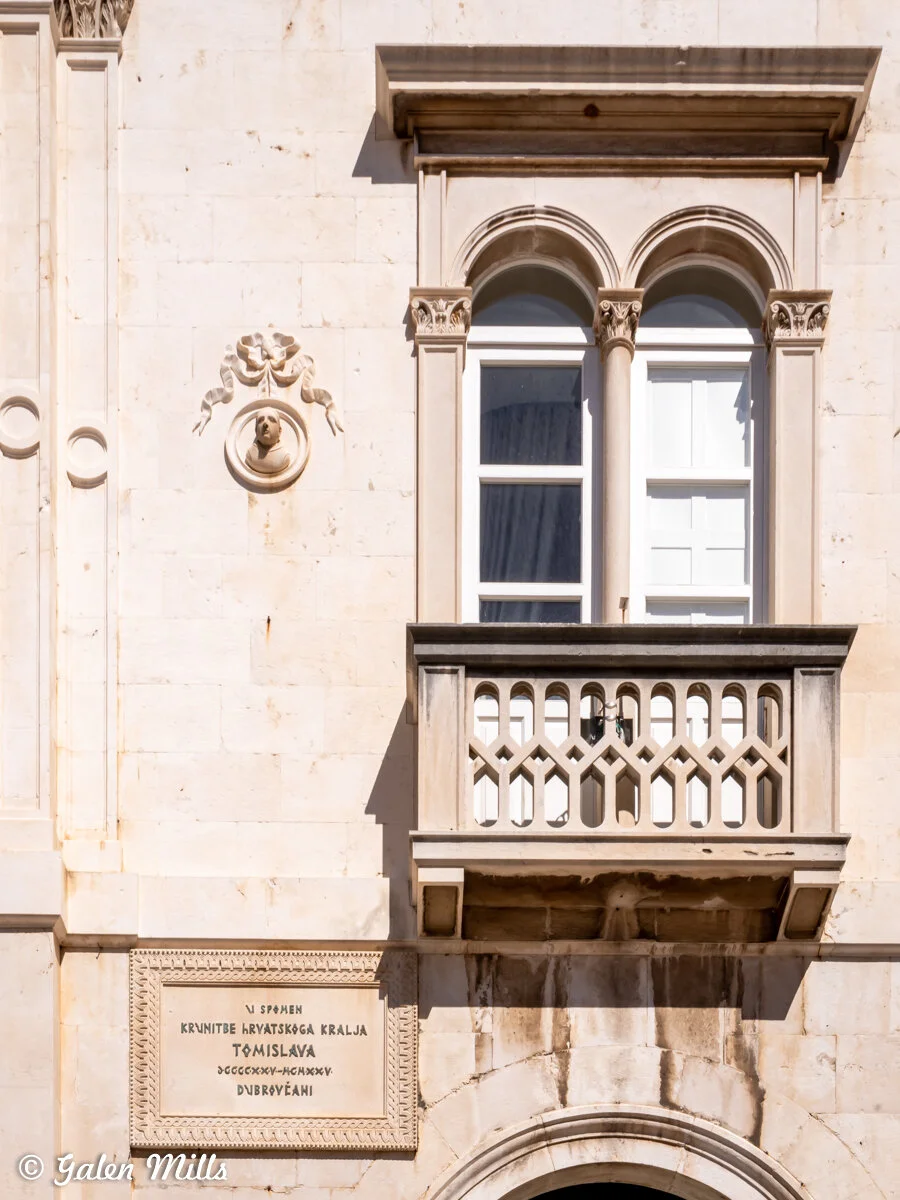 Exterior of a historic building with decorative elements, a double-arched window, a small balcony, and an inscription plaque. The stone facade is light-colored with detailed carvings.