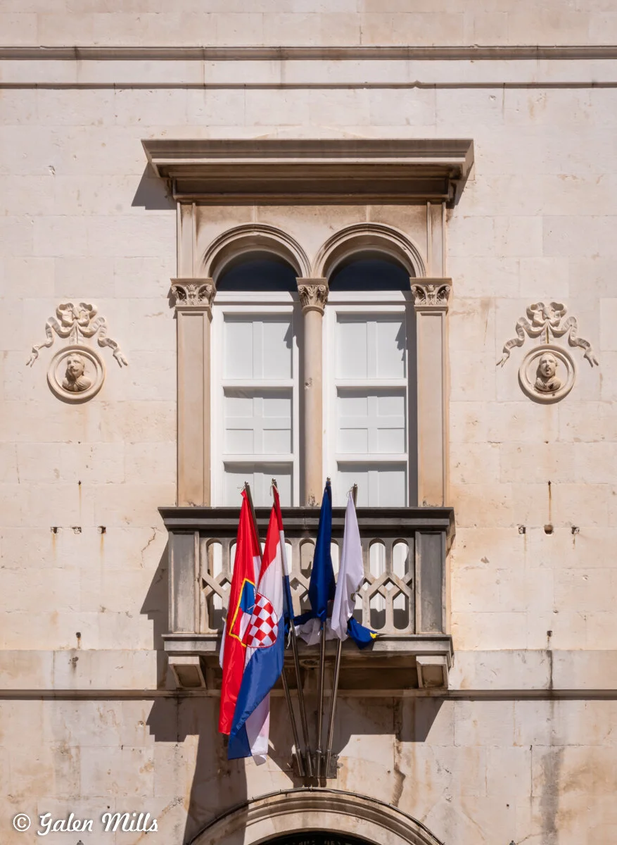 Exterior of a building with a small balcony holding three flags. Two are blue and one is red with a checkered emblem, likely representing Croatia. The building features white double-arched windows and ornate stone carvings.