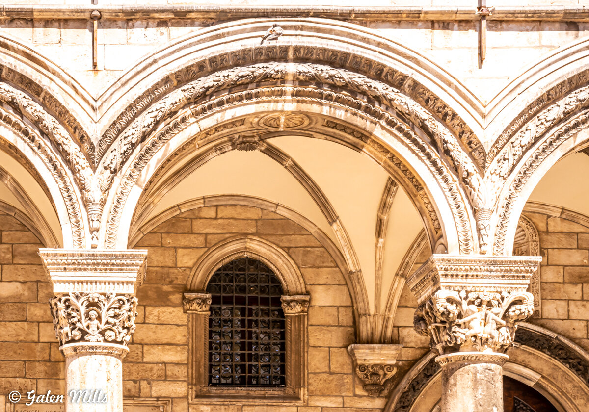 Ornate stone arches and columns with intricate carvings, featuring a recessed window with a grille, typical of historical European architecture.