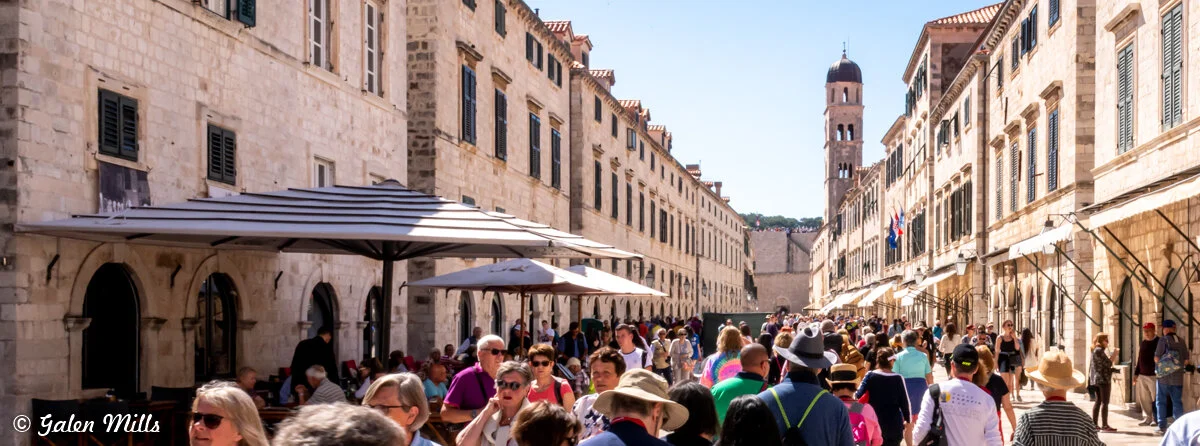 A busy pedestrian street in Dubrovnik with historic stone buildings on either side, and a bell tower in the distance. The street is crowded with people walking, and outdoor cafes are visible under large umbrellas.