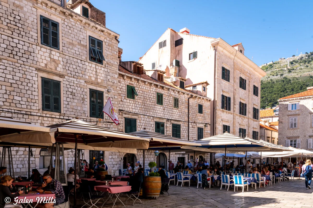 Outdoor café seating with people under umbrellas, stone buildings in the background, and a clear blue sky.
