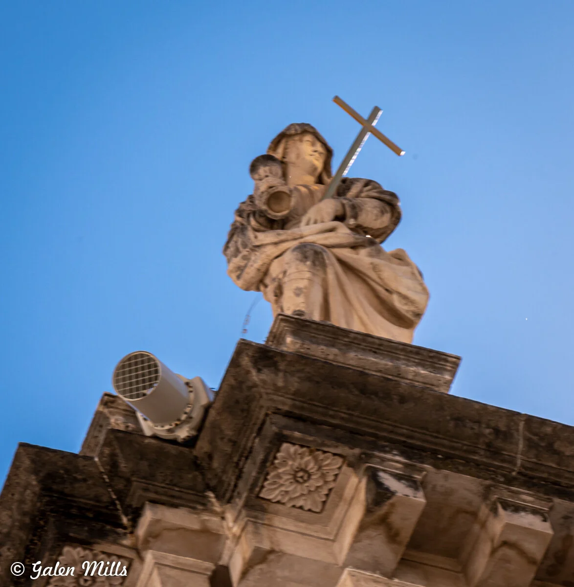 Stone statue of a robed figure holding a cross, set against a clear blue sky, with architectural details and a loudspeaker visible.