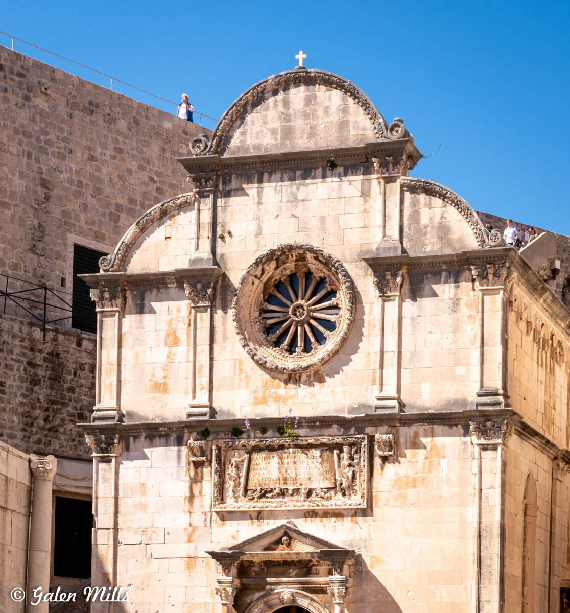 Historic stone building facade with a circular rose window, decorative carvings, and a central arched doorway. A cross is positioned at the peak. Bright blue sky in the background.
