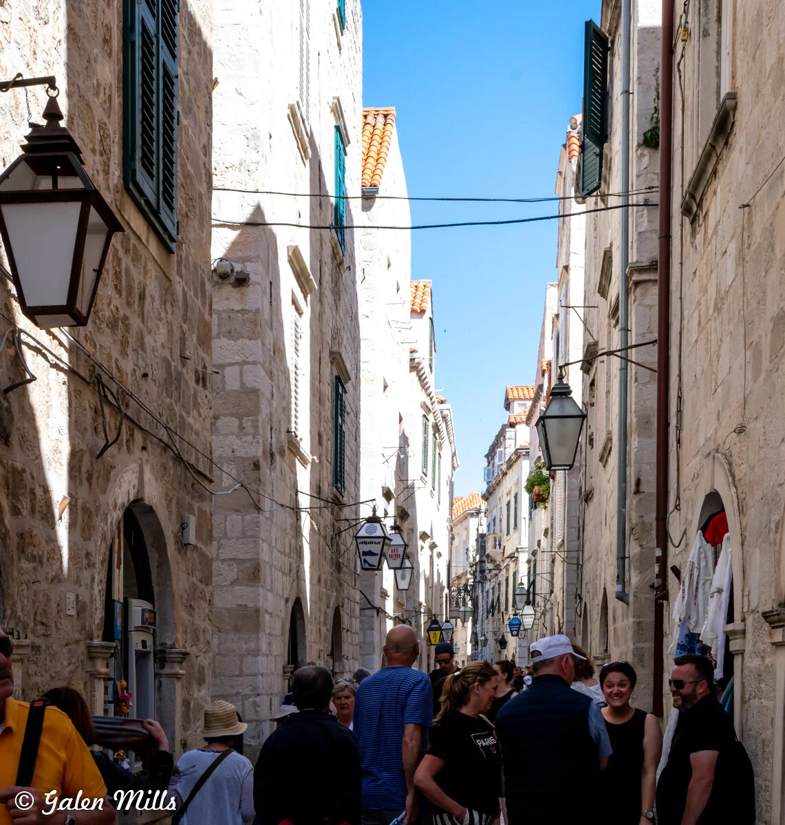 Narrow European street with stone buildings, people walking, and blue sky overhead.