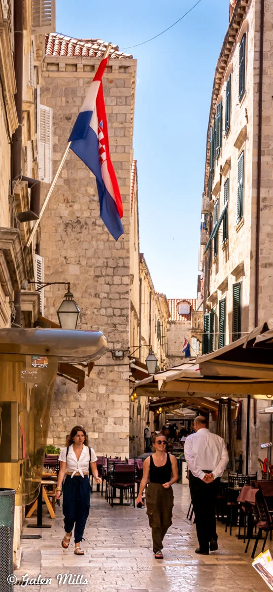 Narrow stone street with Croatian flag, people walking, and outdoor cafes in a European city.